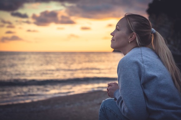 A woman sits on beach and looks at ocean during sunrise.