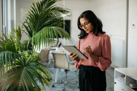 A businessperson in a modern office stands beside a large plant, focused on working with a tablet.