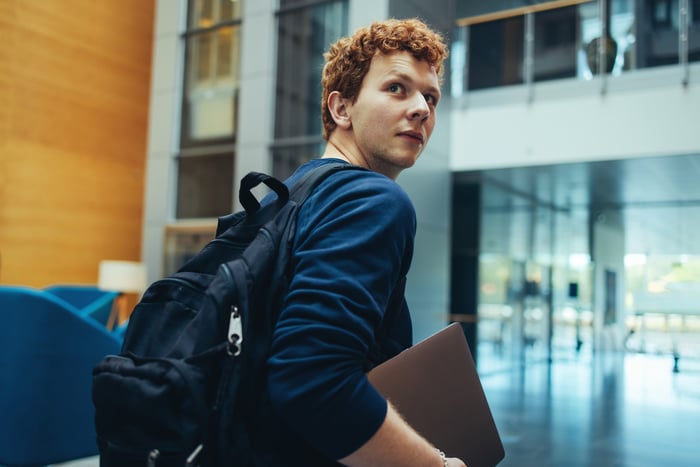 A student holding a laptop and backpack in a school building.