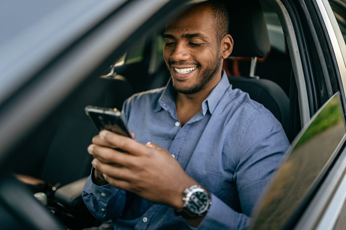 A person in a car smiling while gazing at a smartphone.