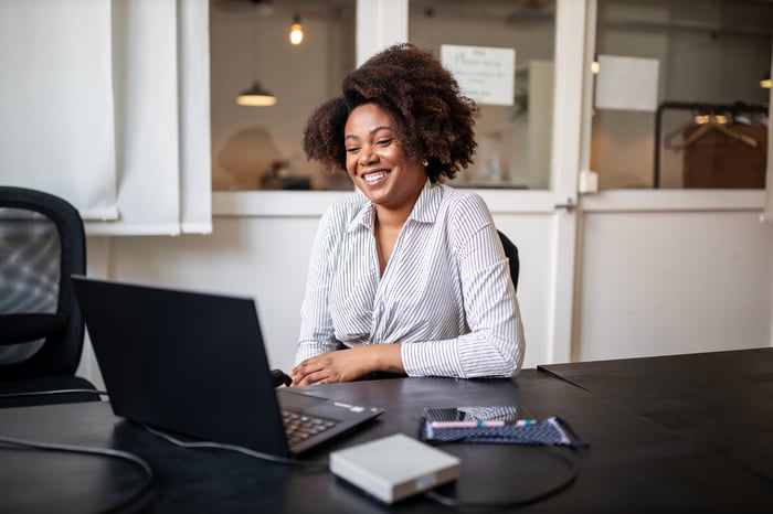 Smiling businessperson is sitting at a desk in an office, engaged on a video call on a laptop.