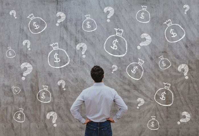 The back of a person who stares at a gray wall of question marks and money bags. 