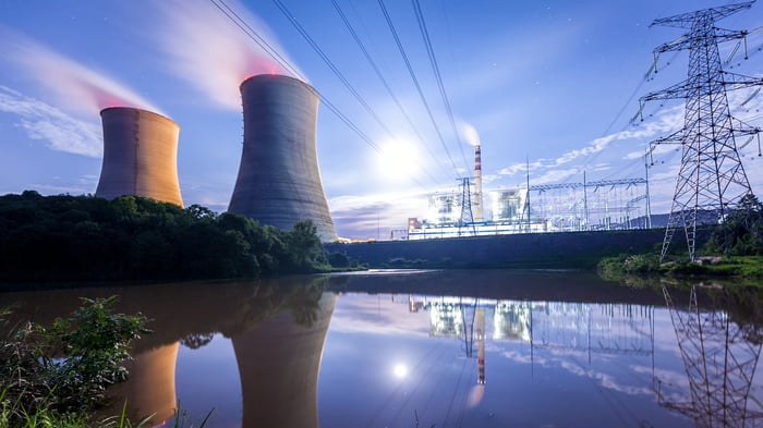 In the background, two nuclear reactors next to power lines. In the foreground a body of water reflects the two reactors. 