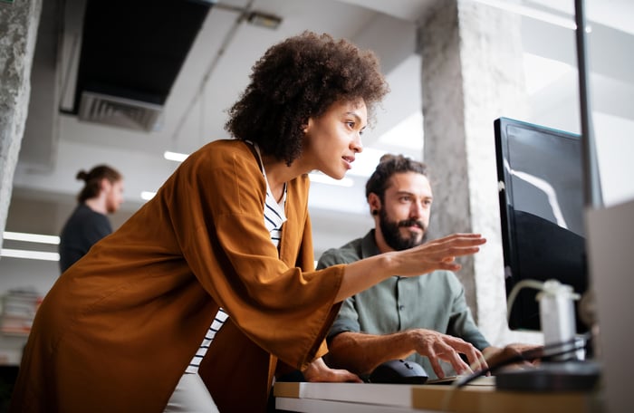 Two office workers discuss content displayed on a computer monitor.