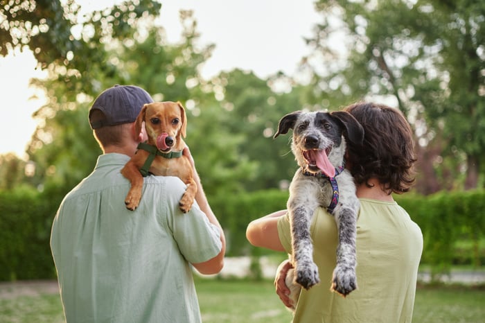 Two people carrying dogs over their shoulders as they walk through a park.