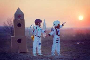 kids dressed as astronauts besides a cardboard rocket.
