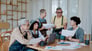 A_group_of_people_discussing_charts_on_paper_while_two_are_reading_on_laptop