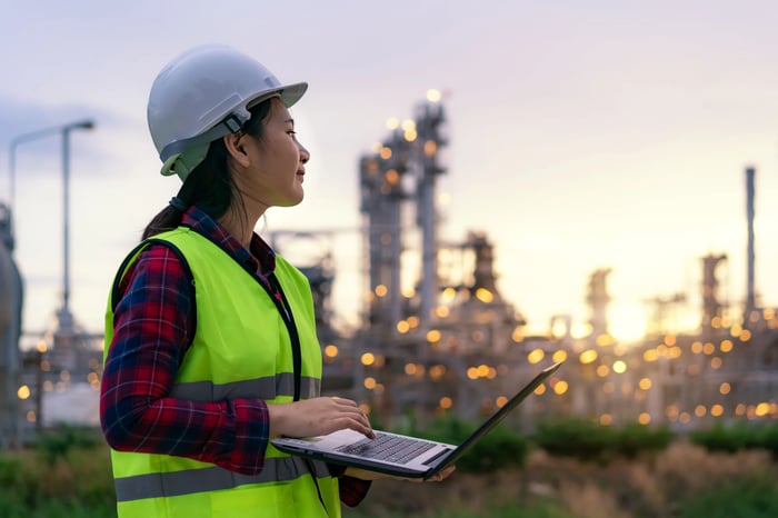 A worker wearing a hard hat and holding a laptop looks out at a power plant in the background. 
