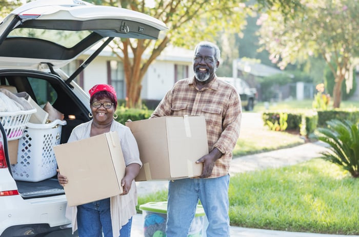 A couple is walking, carrying boxes, and smiling.
