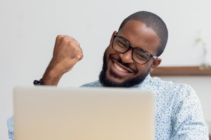 A smiling person holding fist up while looking at a laptop.
