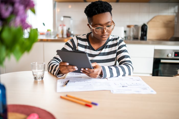 An investor holds a tablet computer as they sit at a table in a kitchen examining some papers.