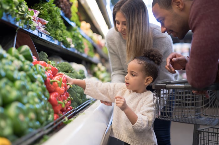 A young child picking out a bell pepper in the produce section, with their parents looking on.