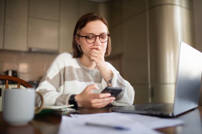 An investor looking at a cell phone while sitting at a table in front of a laptop computer.