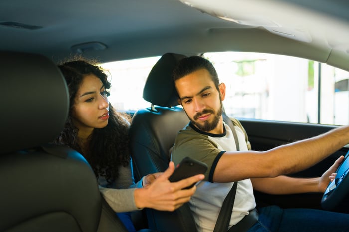 A person in the back seat of a sedan showing the driver directions on a ride-sharing app. 