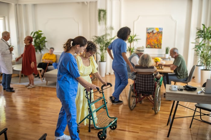 A nursing home common area with many people in it, and a staff member helping a resident in the foreground.