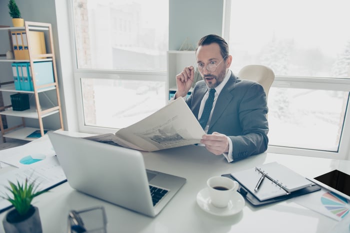 A man in a gray suit wears a shocked expression while he reads a newspaper.