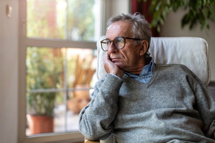 A person sits in a chair at home, looking pensively out a window.