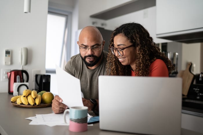 Two investors sit at a kitchen table consulting some papers next to a laptop computer and a mug.