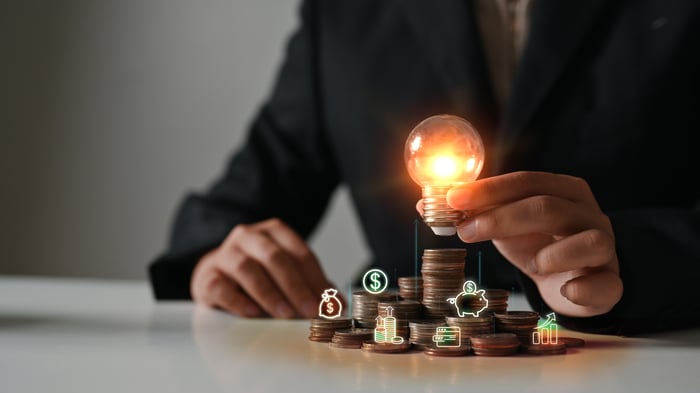 A person holds an illuminated light bulb over stacks of coins.