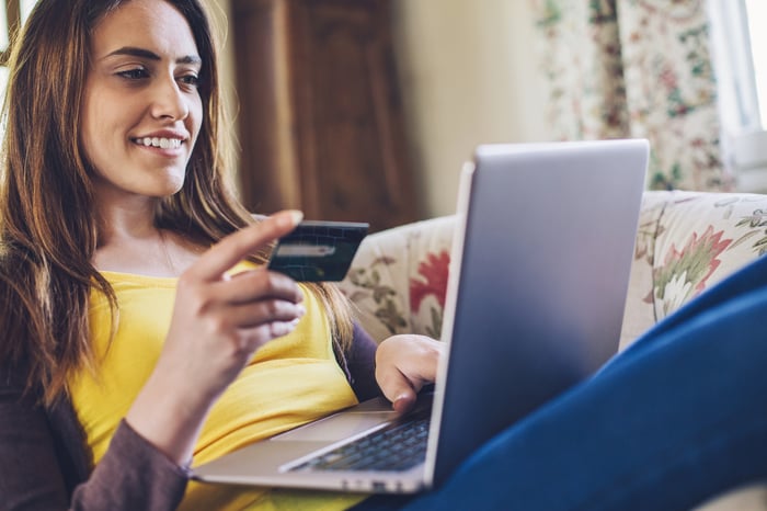 A person smiles while laying on a couch and looking at a laptop computer while holding a payment card.
