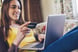 A person smiles while laying on a couch and looking at a laptop computer while holding a payment card.