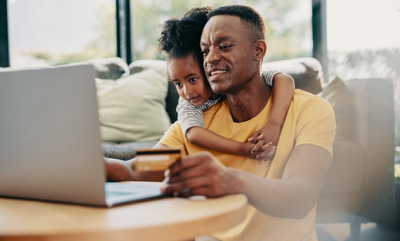 A person smiles while sitting in-front of a laptop computer and holding a payment card while a child is wrapped around their shoulders.