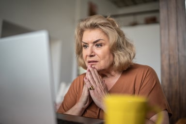 A person clasps their hands and looks at a laptop computer in a concerned manner.