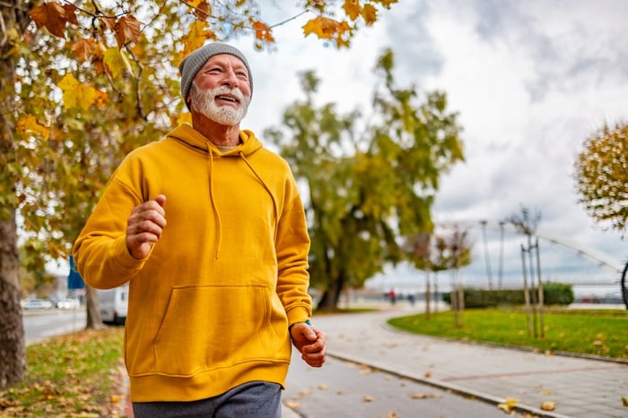 Older person, smiling while jogging down a tree-lined street.