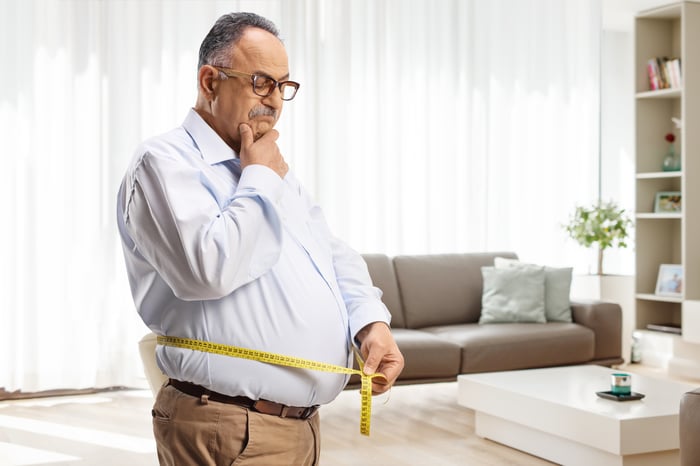 An overweight person looks thoughtfully at the measuring tape they are holding around their waist.