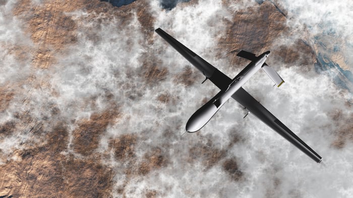 Fixed-wing military drone flying above clouds over barren landscape.