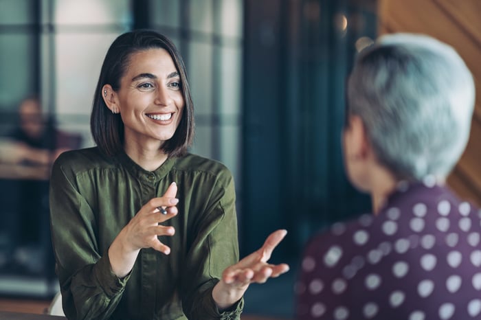 A businessperson is smiling and gesturing with her hands while engaged in a conversation with another person.