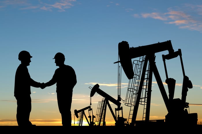 Silhouettes of two people shaking hands next to oil and gas equipment.