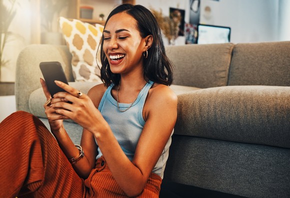 Smiling person sits on floor next to sofa looking at  cellphone.