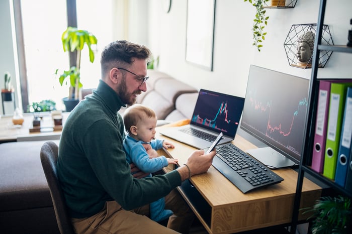 Analyst studying stock charts on laptop and desktop monitor, while checking a smartphone and holding an infant on lap.