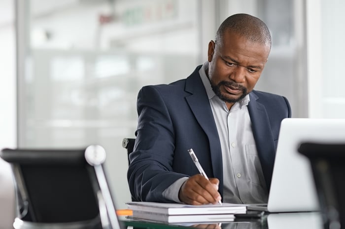An investor scribbling notes into a notebook while seated at a desk in front of a computer.