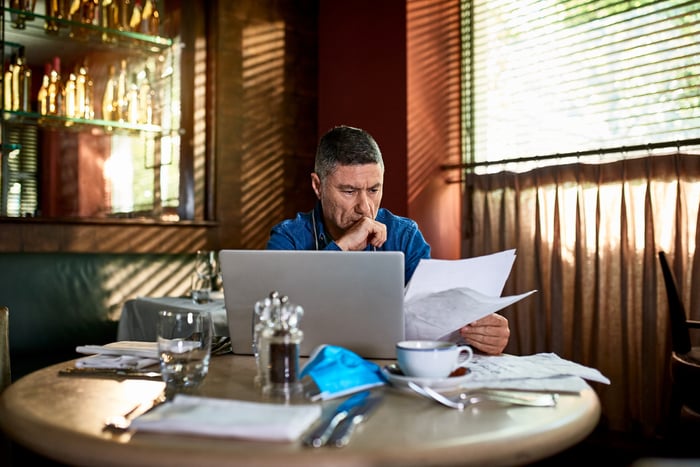 An investor sits in a cafe while looking at some papers and referring to a laptop computer.