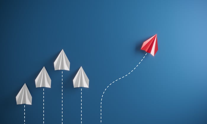 A red paper airplane taking a different path than four white paper airplanes.