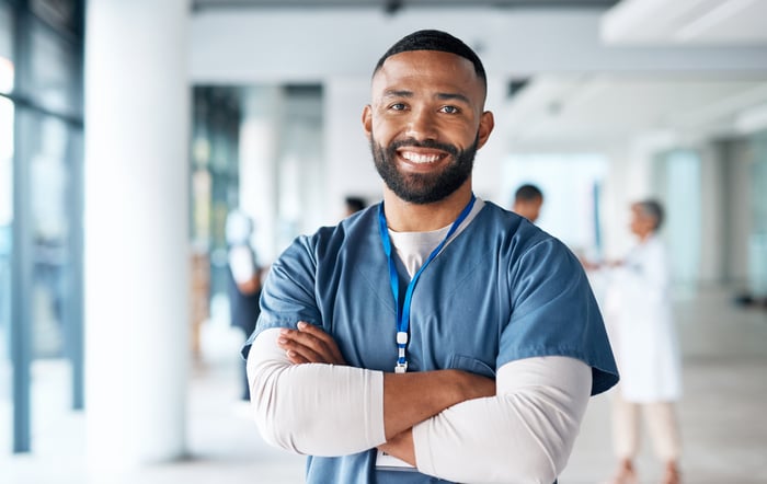 A smiling healthcare professional in scrubs is standing in a medical facility with arms crossed.