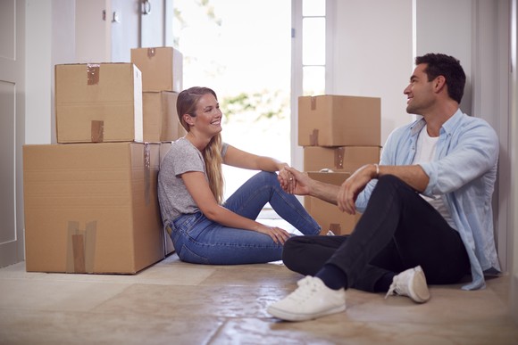 A smiling couple sitting on the floor of their new home, surrounded by boxes.