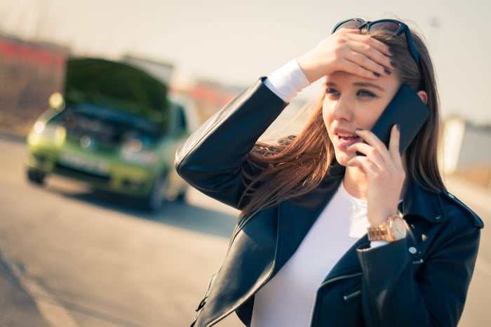 A person talks on a phone in front of a car with its hood up.