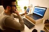 An investor looking at a chart on their computer screen while holding a smartphone