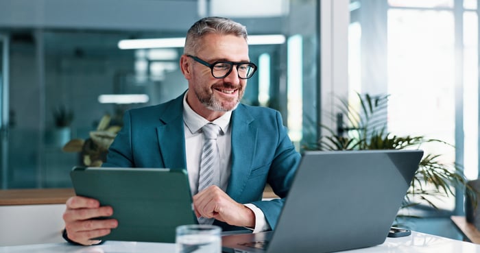 Professional smiling while looking at laptop and holding a tablet in hand.