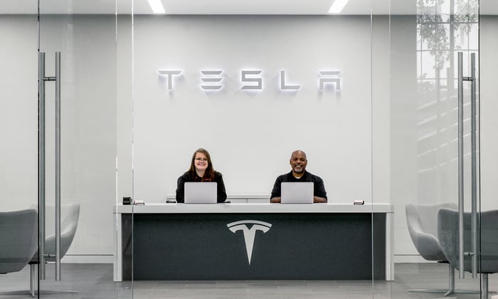 Tesla office with logo over the heads of two employees sitting in front of laptops.