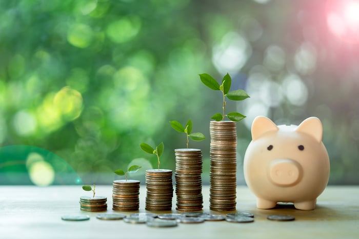 Plants sprout from stacks of coins next to a piggy bank.