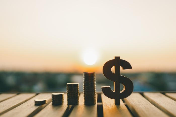 The sun is shining on stacks of coins rising in a stairstep manner next to a dollar symbol.