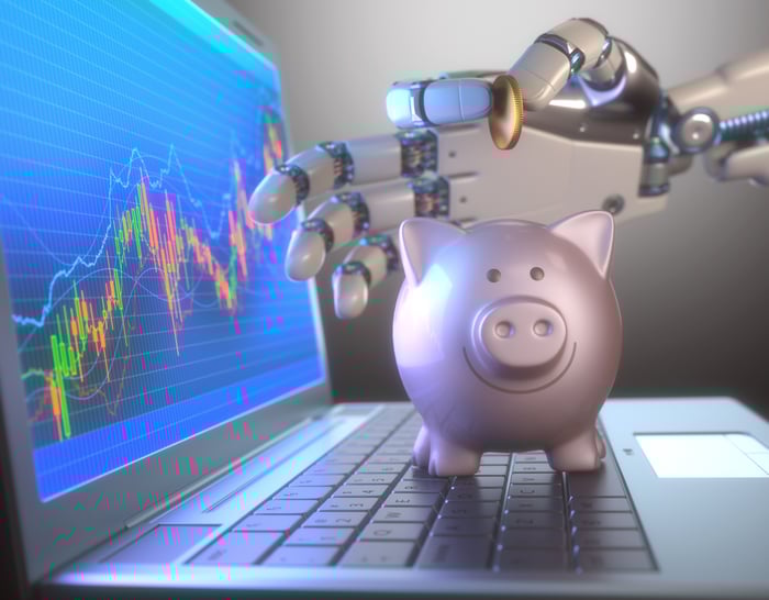 A robotic hand holds a coin above a piggy bank that is standing on the keyboard of a laptop.