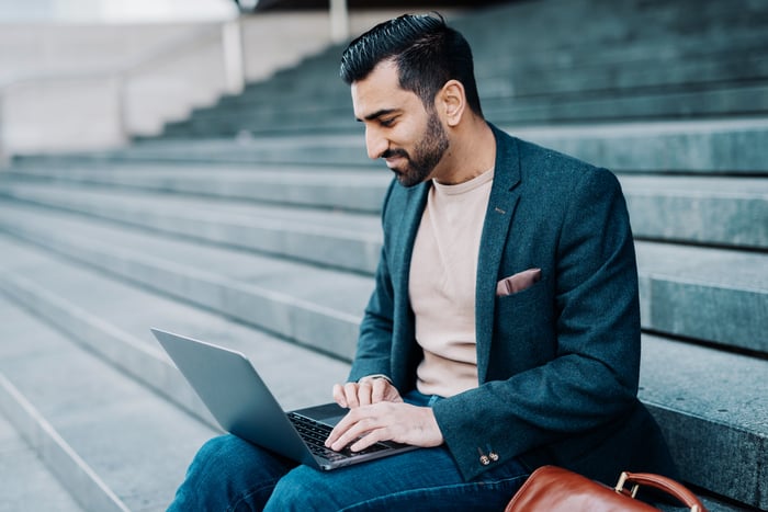 An investor works on a laptop computer while sitting on steps in a public setting.