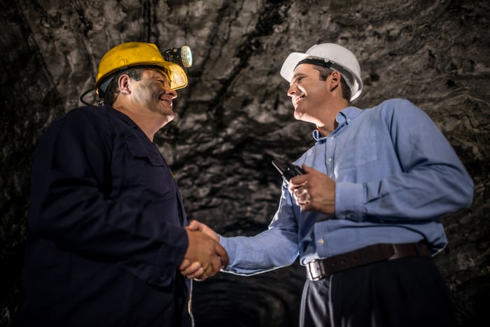 Two people wearing hard hats are smiling and shaking hands while inside a mine.