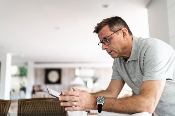 An investor looks exasperated while leaning on a kitchen counter while consulting a tablet computer.