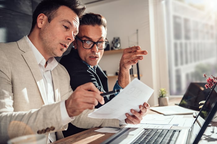 Two investors sitting in an office in front of two laptops consult some papers and discuss them.